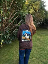 Remera de algodón color marrón, de manga larga y corte holgado. Presenta una estampa rectangular en la espalda con un diseño de un caballo, montañas y el texto 'Sierra Morena Uruguay'.