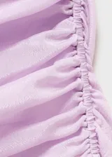 Vestido corto de satén en color lila, con diseño entallado y cuello asimétrico. Presenta tirantes finos, detalle de fruncido lateral con lazo ajustable y una pequeña abertura en el bajo. Cuenta con espalda abierta y forro interior.