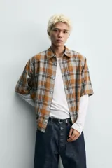 Camisa de corte boxy fit confeccionada en tejido de algodón, con estampado de cuadros en tonos tierra. Presenta cuello solapa, manga corta y cierre frontal de botones.