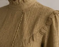 Blusa color mostaza con textura de pequeños puntos y detalles calados verticales. Presenta cuello alto con volados, volados decorativos en los hombros y mangas largas con puños elastizados.