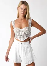 Top tipo corset de encaje blanco con diseño de copas estructuradas, tirantes anchos con borde festoneado y cierre frontal con cordones ajustables.