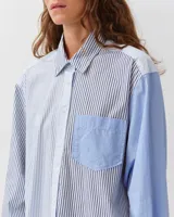 Camisa de corte recto y ligeramente oversize, confeccionada en algodón orgánico. Presenta un diseño a rayas con paneles en contraste de tonos azules y blancos, cuello clásico, cierre frontal de botones y un bolsillo en el pecho.