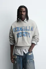 Sudadera gris de corte boxy fit, con cuello redondo y manga larga. Presenta un estampado frontal con texto en azul con efecto desgastado y acabados en rib en puños y ruedo.