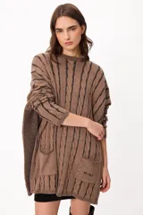 Sweater de punto con diseño de trenzas verticales, color crudo y camel, con bolsillos frontales y detalle de logo bordado. Presenta un corte holgado y mangas largas.