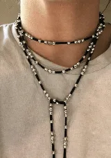 Corbatín largo de cordón negro con cuentas plateadas distribuidas a lo largo de toda la pieza. Su diseño versátil permite usarlo con múltiples vueltas alrededor del cuello o como un collar tipo lariat ajustable.