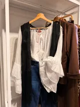Blusa romántica de color blanco, confeccionada en tejido ligero y semitransparente, con escote redondo fruncido y cordones ajustables con tiras colgantes. Presenta mangas largas amplias de diseño fluido.