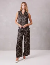 Conjunto de dos piezas compuesto por una blusa sin mangas con cuello solapa y escote en V con botones frontales, y un pantalón de tiro alto con pierna ancha y bolsillos laterales. Ambas prendas están confeccionadas en satén con un estampado floral sobre fondo oscuro.