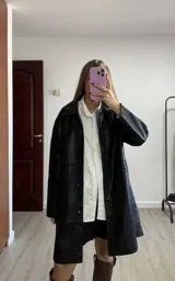 Trench de corte recto confeccionado en eco cuero negro, con cuello clásico, cierre frontal con botones y forro interior completo.