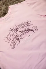 Remera de algodón elastizado color rosa pálido, con cuello redondo y manga corta. Estampado frontal en tonos morados con texto que dice "ARTE INMORTAL CUERPO MORTAL" y un diseño de relámpago.