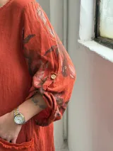 Vestido largo color terracota con diseño asimétrico que incluye un lateral con estampado floral grande. Presenta mangas tres cuartos, escote redondo y un bolsillo aplicado en el frente.