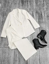 Conjunto sastrero de dos piezas color crudo, compuesto por un blazer cruzado de doble botonadura y bolsillos con solapa, y un pantalón de tiro alto con corte recto y pinzas en la cintura.