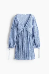 Vestido corto de punto plisado con escote en V pronunciado, costuras diagonales bajo el busto y mangas globo largas con tiras ajustables en los puños. Presenta un diseño evasé con estampado floral en tonos azules sobre fondo blanco.
