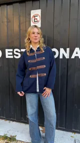Campera de estilo militar en color azul marino, confeccionada en tejido de punto grueso. Presenta un diseño de cuello redondo, cierre frontal con tiras de cuero marrón y botones metálicos decorativos. Posee un corte recto y holgado.