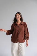 Camisa de color marrón chocolate, con cuello clásico, cierre frontal de botones y mangas largas abullonadas con detalle de tejido calado en los puños.