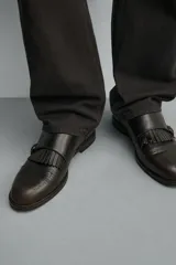 Zapato de vestir estilo monk strap fabricado en piel color marrón oscuro. Presenta ajuste mediante dos hebillas laterales metálicas, detalle de flecos en el empeine y puntera con perforaciones decorativas tipo brogue.