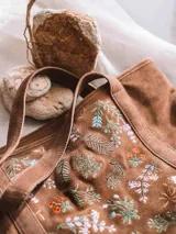 Bolso tipo tote bag confeccionado en cuero serraje color marrón, con diseño artesanal que incluye bordados florales coloridos en el panel frontal. Cuenta con asas de hombro y cierre superior.