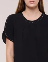 Blusa holgada de color negro, con escote redondo y mangas cortas adornadas con apliques de tachas metálicas. Presenta delicadas tablas centrales en el frente y un diseño elegante.