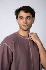 Remera de corte oversize color malva, confeccionada en algodón pesado, con costuras externas visibles en color blanco que recorren el centro, el cuello y los bordes de las mangas.