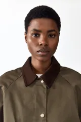 Campera de sarga color verde militar, con cuello camisero de pana marrón oscuro, cierre frontal con solapa y botones a presión metálicos, bolsillos frontales grandes con solapa y mangas raglán.