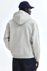Campera con capucha de felpa gris melange, con cierre frontal, bolsillos tipo canguro y cordón ajustable. Presenta hombros caídos, corte holgado y puños y bajo en punto acanalado.
