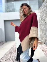 Poncho tejido de punto con diseño de bloques de color en tonos bordó, beige, blanco y negro, con terminación de flecos en el ruedo.