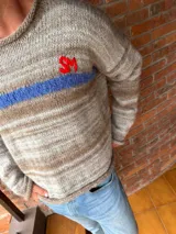 Sweater de tejido de punto con efecto jaspeado en tonos beige y marrón. Presenta una franja horizontal azul brillante a la altura del pecho con las iniciales 'SM' bordadas en rojo. Tiene cuello redondo y puños y ruedo en punto elástico.