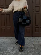 Cartera de hombro confeccionada en cuero y gamuza con diseño de damero, combinando paneles de texturas lisas y aterciopeladas. Presenta una manija trenzada y detalles de costuras artesanales en los laterales.