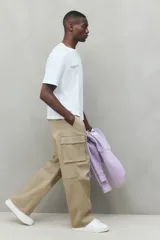 Pantalón cargo de corte holgado en color beige, confeccionado en tejido de algodón. Presenta cintura elástica con cierre de botón a presión, bolsillos laterales al bies, bolsillos de fuelle con solapa en las piernas y pliegues decorativos en las rodillas.