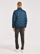Chaqueta acolchada de peso medio en color azul petróleo, con cierre frontal de cremallera, cuello alto y parche de silicona con el logotipo de la marca en el pecho. Cuenta con bolsillos laterales y ribetes elásticos en puños y dobladillo.