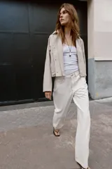 Trench corta de color beige, con cuello solapa, mangas largas con trabillas en los hombros, bolsillos delanteros de vivo y cierre frontal cruzado con botones a presión.