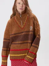 Pullover de punto con textura aterciopelada, diseño oversize, cuello polo con cierre frontal parcial y detalles de pespuntes en contraste. Presenta rayas horizontales en tonos tierra y un parche bordado con la letra M en el pecho.