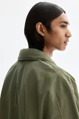 Campera estilo cargo de tejido ripstop, color verde militar, con cuello elevado que incluye capucha oculta. Presenta cierre frontal con solapa rompevientos, cuatro bolsillos delanteros con solapa y botón a presión, puños ajustables y cordón interno en la cintura.