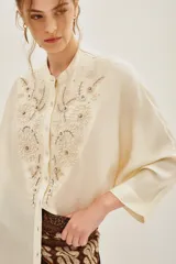 Camisa de viscosa color crema, con cuello mao y cierre frontal de botones. Presenta un delicado bordado floral en el frente y mangas tres cuartos con puños rectos.