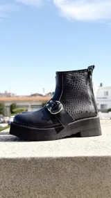Botas cortas negras con plataforma y suela robusta. Presentan una combinación de texturas en cuero liso y acabado con textura tipo reptil en la caña. Cuentan con una tira con argolla metálica decorativa en el lateral y cierre trasero.