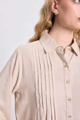 Vestido camisero corto de color beige, con cuello clásico, cierre frontal de botones y detalle de tablas verticales en el frente. Presenta mangas tres cuartos y un corte holgado con ruedo redondeado.