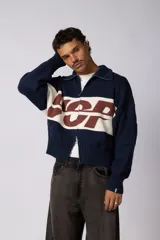 Campera tejida de punto con cierre frontal completo y cuello polo. Presenta un diseño con franja horizontal en color azul y letras en blanco sobre fondo negro, con puños y ruedo acanalados.