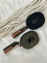 Monedero circular de color negro con cierre perimetral. Presenta una etiqueta frontal con el logo de la marca y una correa lateral de tela con franjas en tonos beige, marrón y negro.