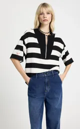 Blusa de punto con estampado de rayas horizontales en blanco y negro. Presenta un corte holgado, cuello redondo con panel frontal sólido en contraste, cierre de cremallera metálica, mangas cortas anchas y un parche bordado en la manga derecha.