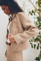 Chaqueta corta de tejido texturizado en color tostado, con cuello camisero, cierre frontal de botones metálicos dorados y diseño entallado con costuras moldeadoras.
