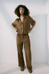 Pantalón de tiro medio color marrón, con cintura elástica y cordón ajustable con abalorios decorativos. Presenta bolsillos laterales y perneras rectas con diseño texturizado.