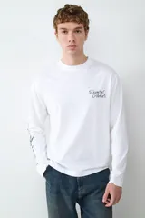 Camiseta de manga larga color blanco, con cuello redondo y corte holgado. Presenta un estampado tipográfico en el pecho y detalles gráficos en la manga.