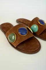Sandalias planas de gamuza marrón con tira ancha decorada con dos apliques circulares de piedras en tonos verde y azul, enmarcados en metal dorado.