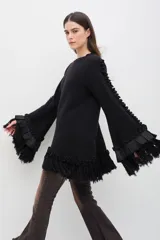 Sweater de lana merino color beige, con silueta amplia y relajada. Presenta mangas largas con volumen, terminaciones con volados de lino y flecos en los puños y en el ruedo, además de detalles trenzados a lo largo de las mangas.