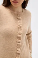 Saco de punto color gris, con cuello redondo, cierre frontal con botones y detalle de volados en la tapeta y el ruedo. Presenta mangas largas con puños acampanados.