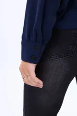 Camisa blanca de manga larga con cuello clásico y cierre frontal de botones negros en contraste. Presenta un bolsillo frontal en el pecho y un diseño de líneas simples y corte recto.