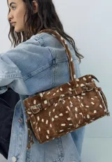 Cartera de tamaño medio confeccionada en cuero vacuno con textura de pelo y estampado animal print tipo vaca. Presenta estructura rígida, doble asa de hombro, cierre superior, detalle de tira horizontal con tachas y hebillas metálicas, y lazos decorativos en los laterales.