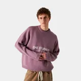 Sweater de tejido punto color rosa, con cuello redondo y corte oversize. Presenta un estampado frontal con el texto "EME STUDIOS" y debajo "curated by ben ice".