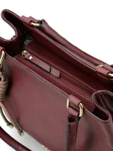 Cartera de hombro color bordó con textura granulada, diseño estructurado y herrajes dorados. Incluye un detalle decorativo de nudo en tono beige claro y logo metálico en la parte frontal.