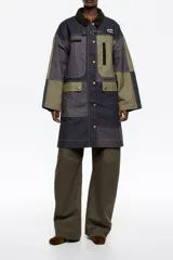 Gabardina midi de algodón con diseño patchwork en tonos azul marino, gris y verde oliva. Presenta cuello de pana, cierre frontal con botones a presión y gancho metálico, bolsillos laterales con solapa y costuras a la vista.