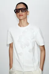 Camiseta blanca de algodón con cuello redondo y manga corta, destacada por aplicación de flores tridimensionales a tono en el frente.